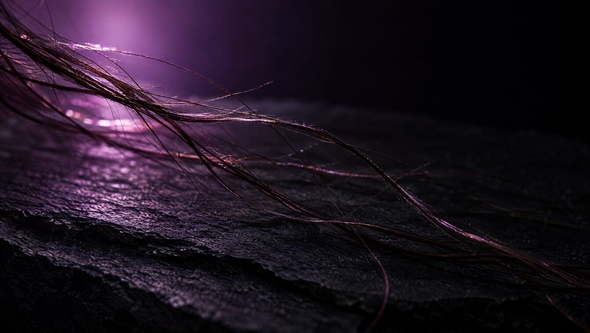 Loose strands of dark hair trailing across a dark slate surface, backlit by a soft plum light.