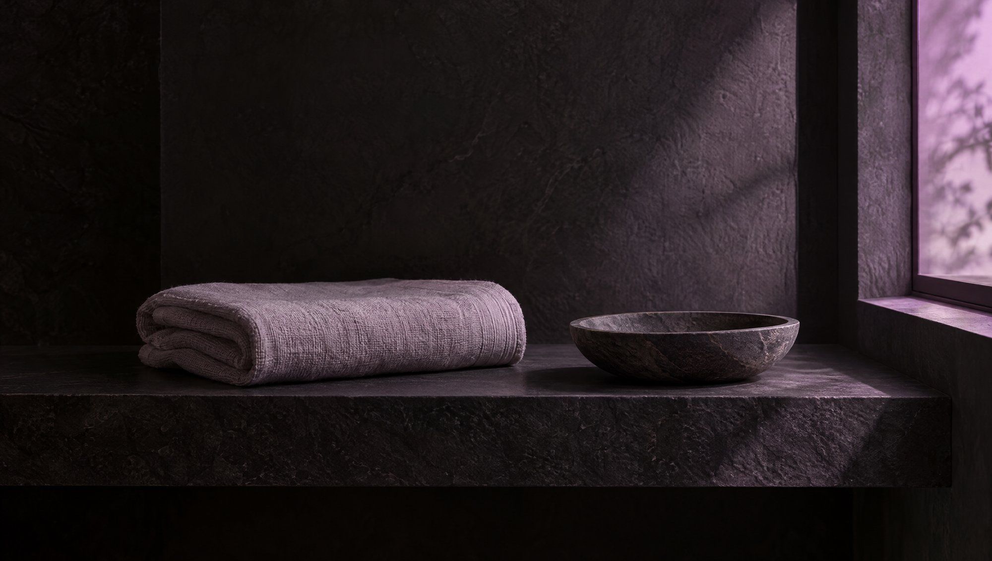 A folded linen towel beside an empty stone dish on a dark bathroom shelf, lit by plum window light.