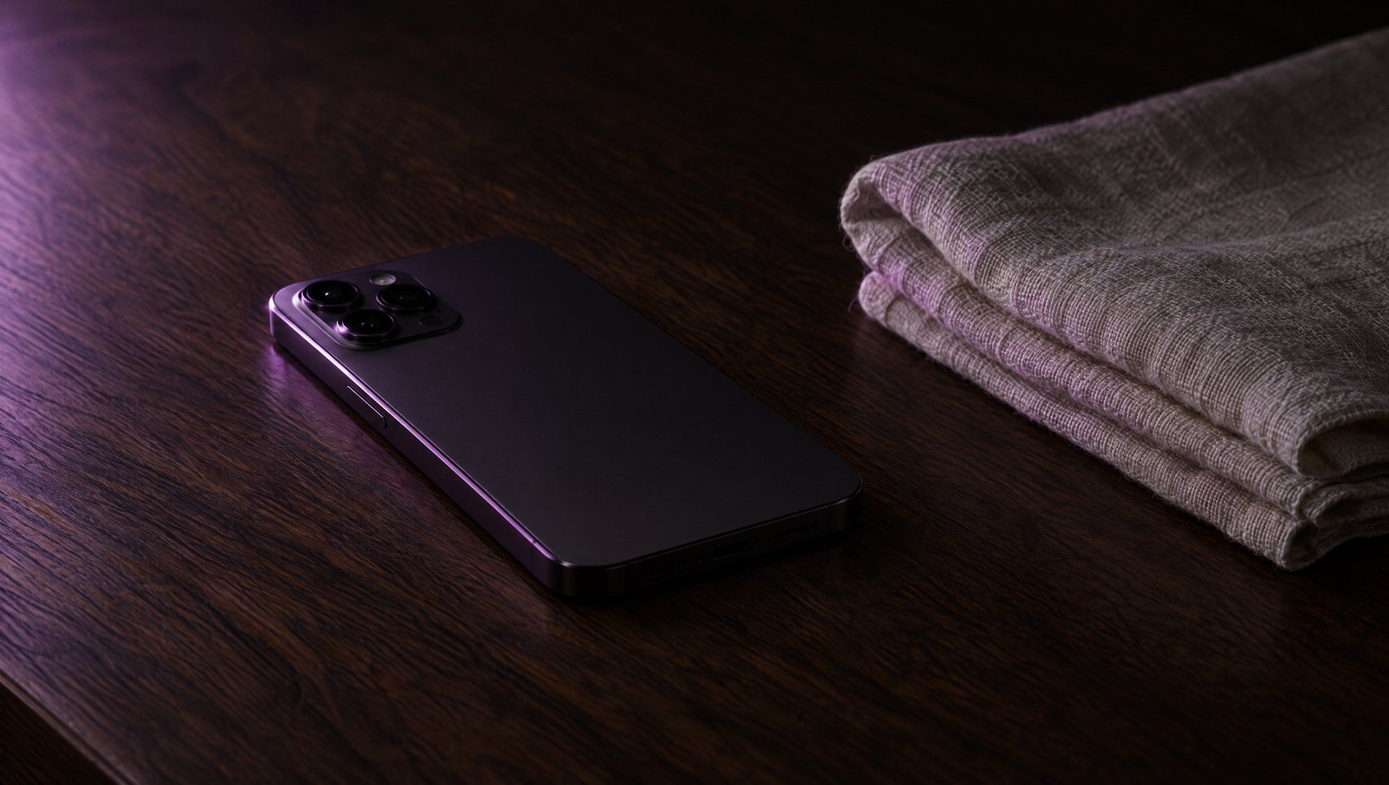 A smartphone lying face down on a dark wooden table beside a folded linen towel, its edges catching plum side light.