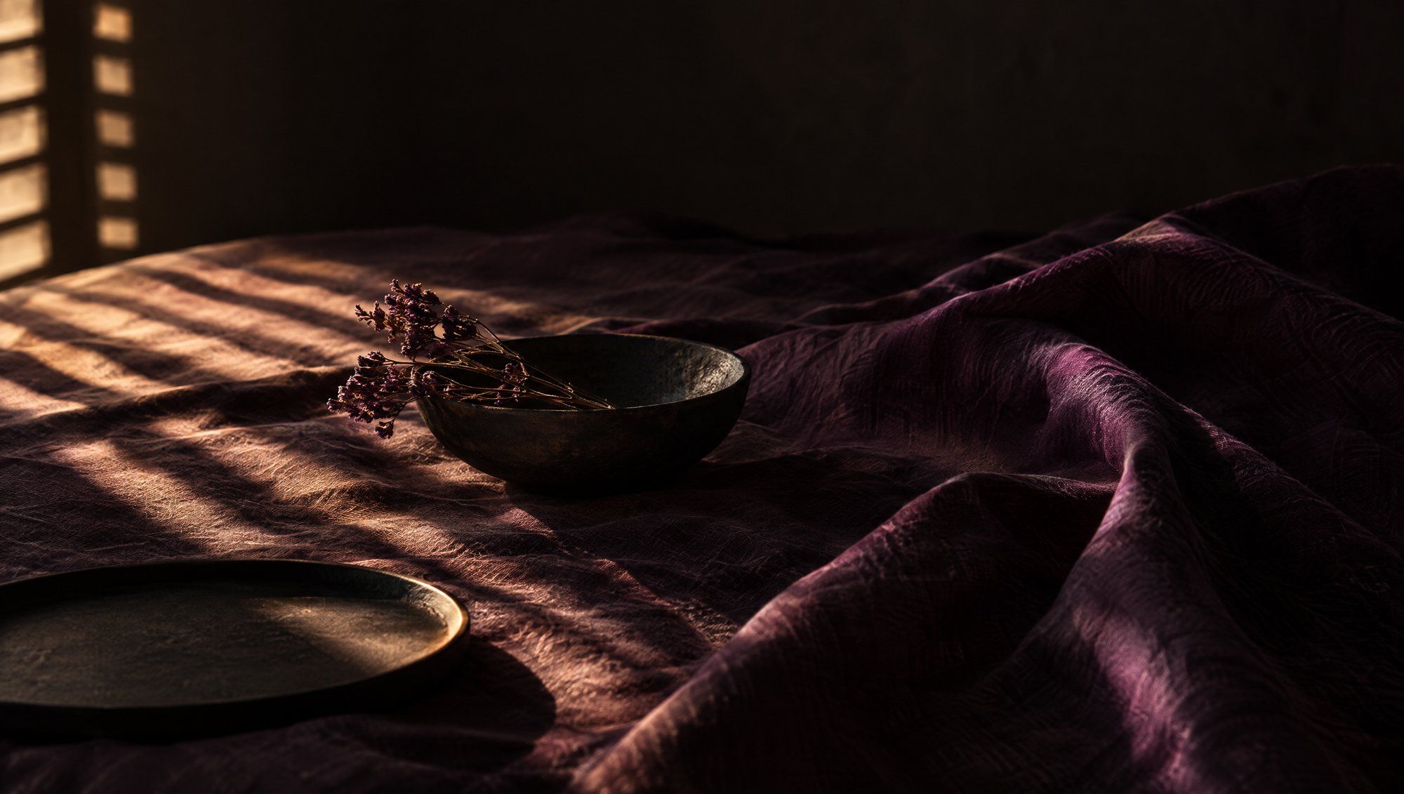 Bands of sunlight from window slats falling across rumpled dark plum linen in a dim room, with a dark stone bowl, a sprig of dried flowers and a flat dark plate.